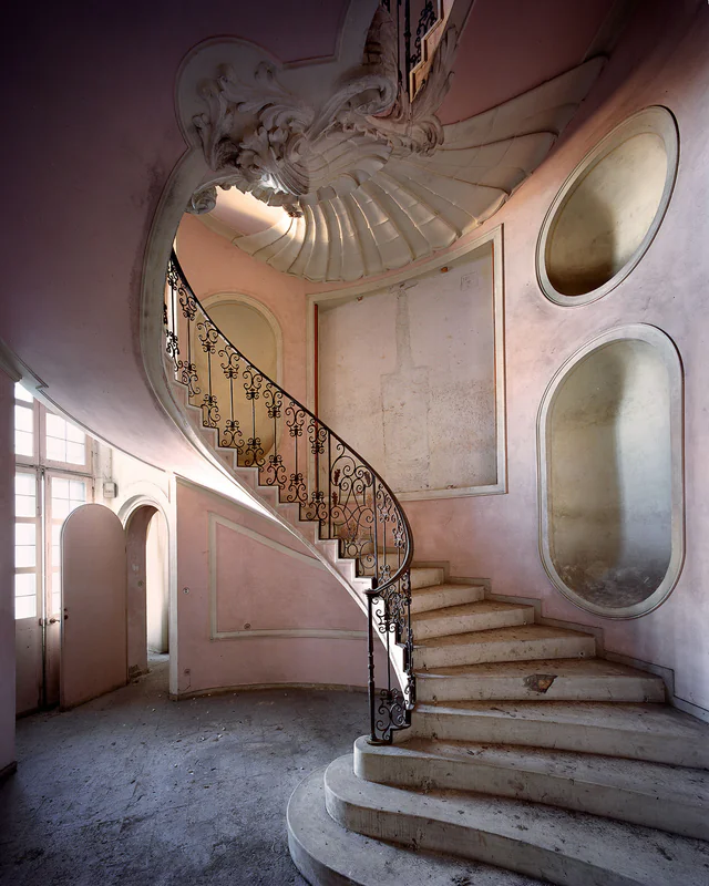 A spiral staircase with a wrought-iron railing in the pink stairwell of an abandoned palace
